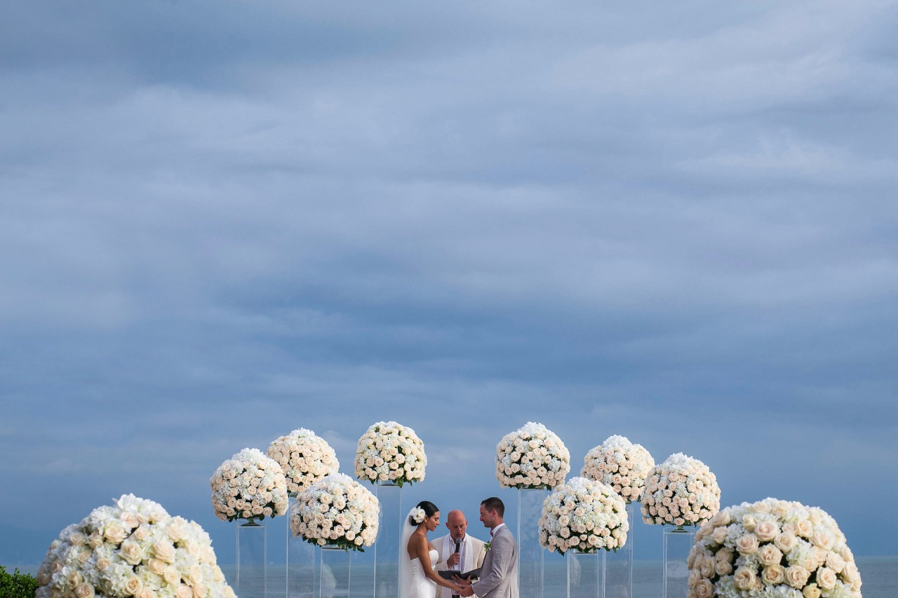 A bride, groom and priest in front of white flower arragement in the beach